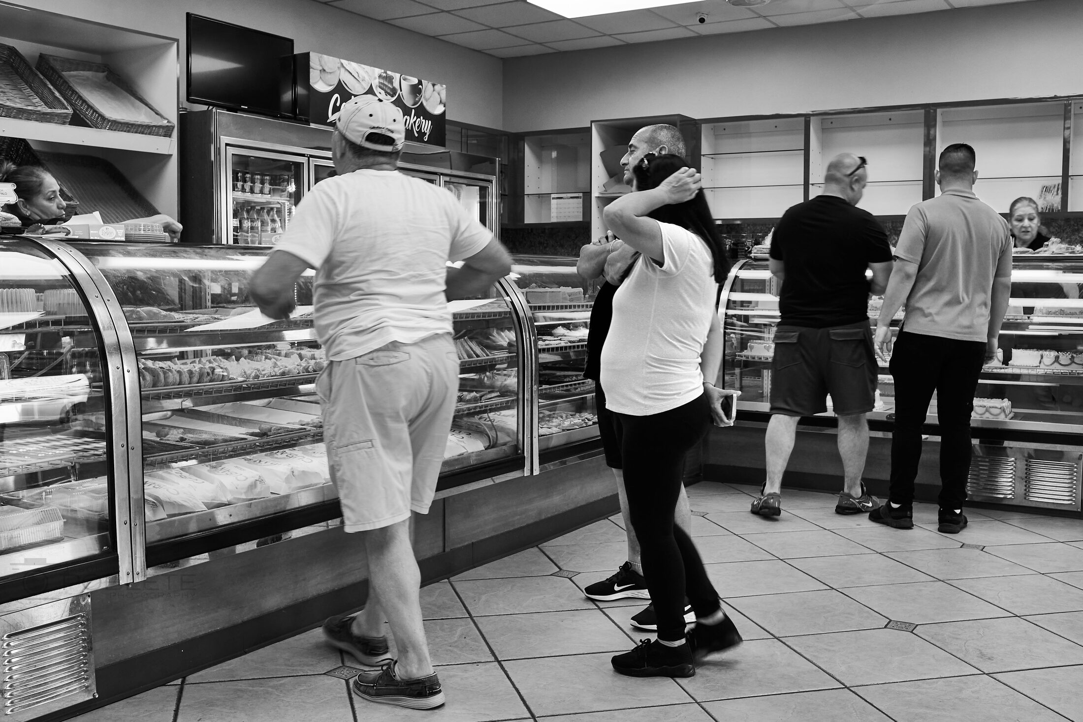 Customers wait at a bakery counter beneath display cases.