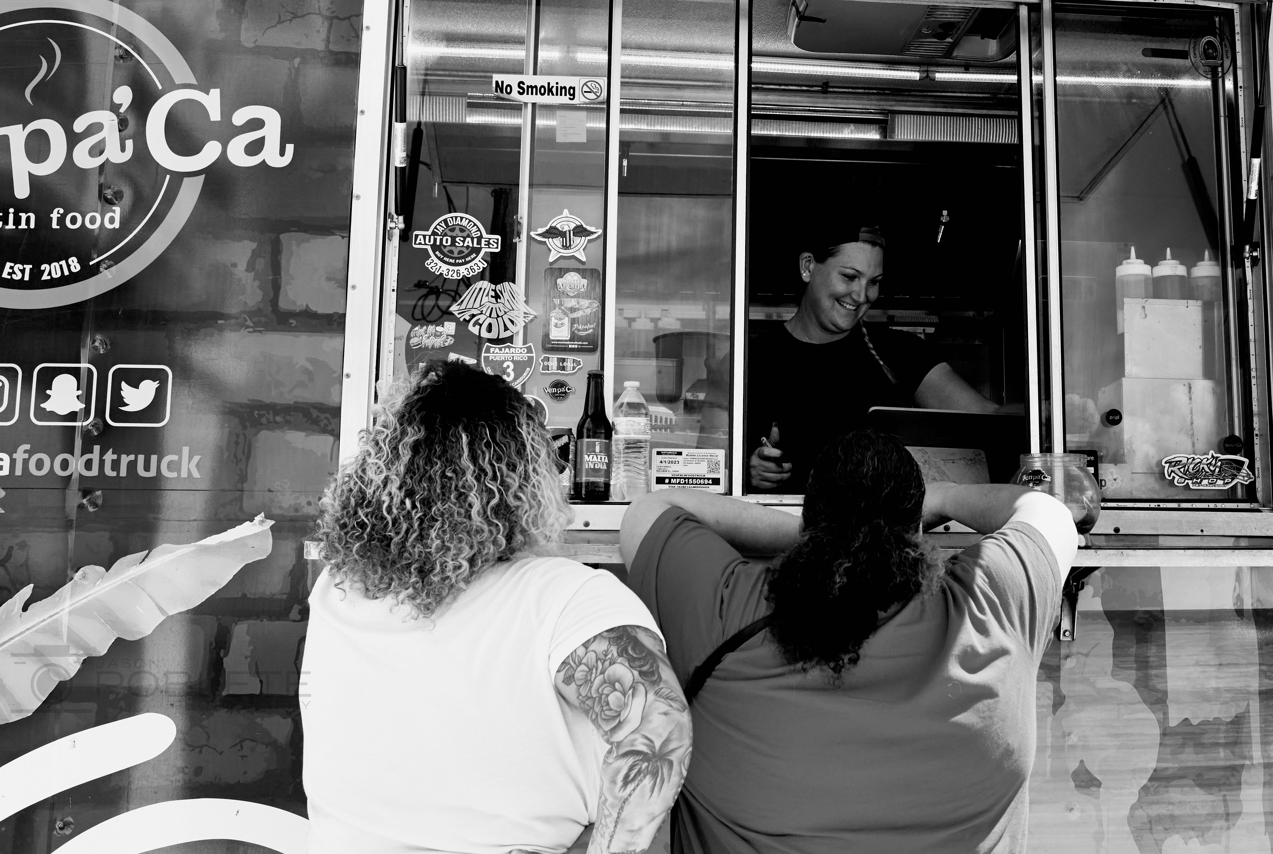 Two customers order food from a smiling worker at a food truck window.