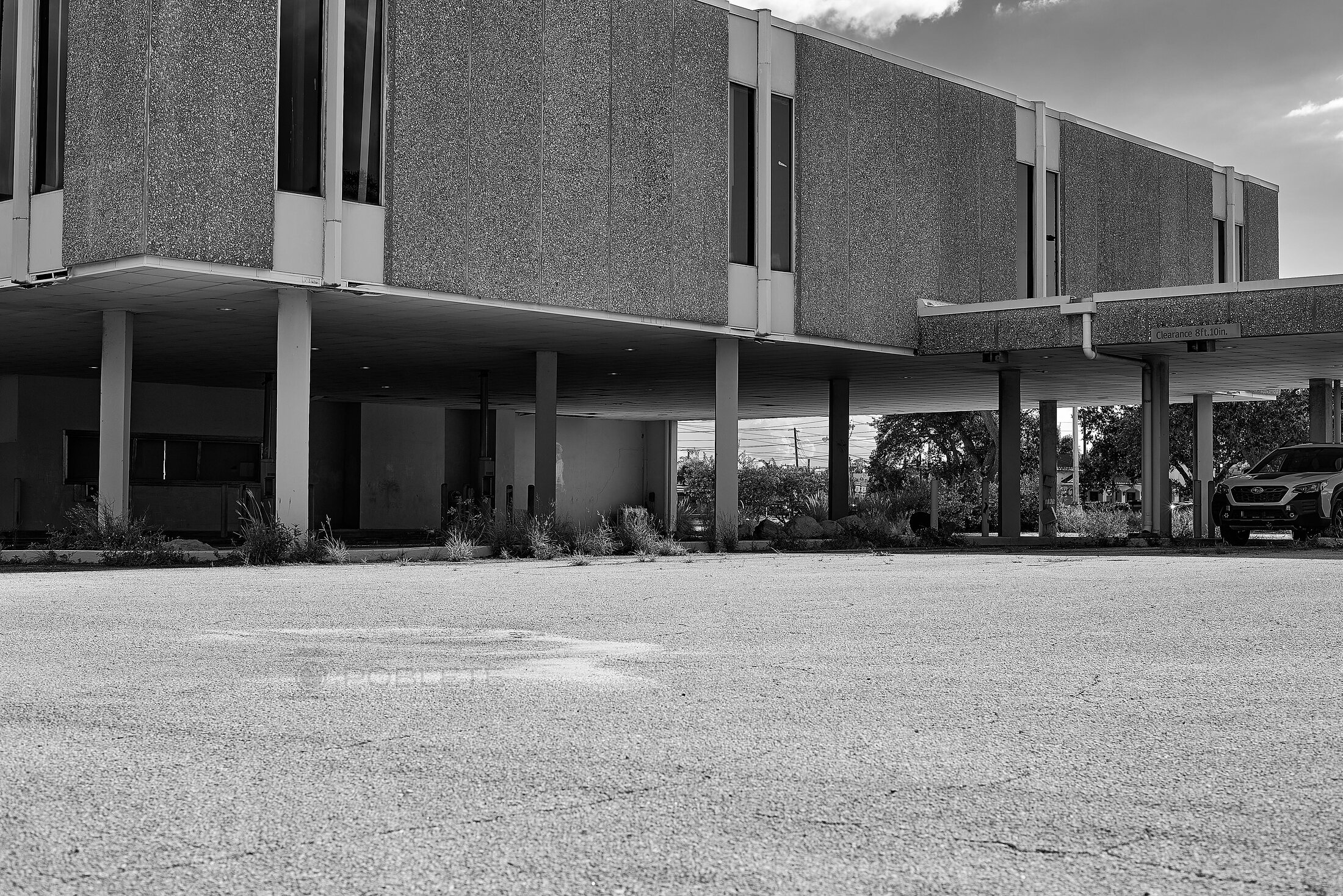 A vacant commercial building stands above an empty parking lot, with weeds growing around its covered entrance.