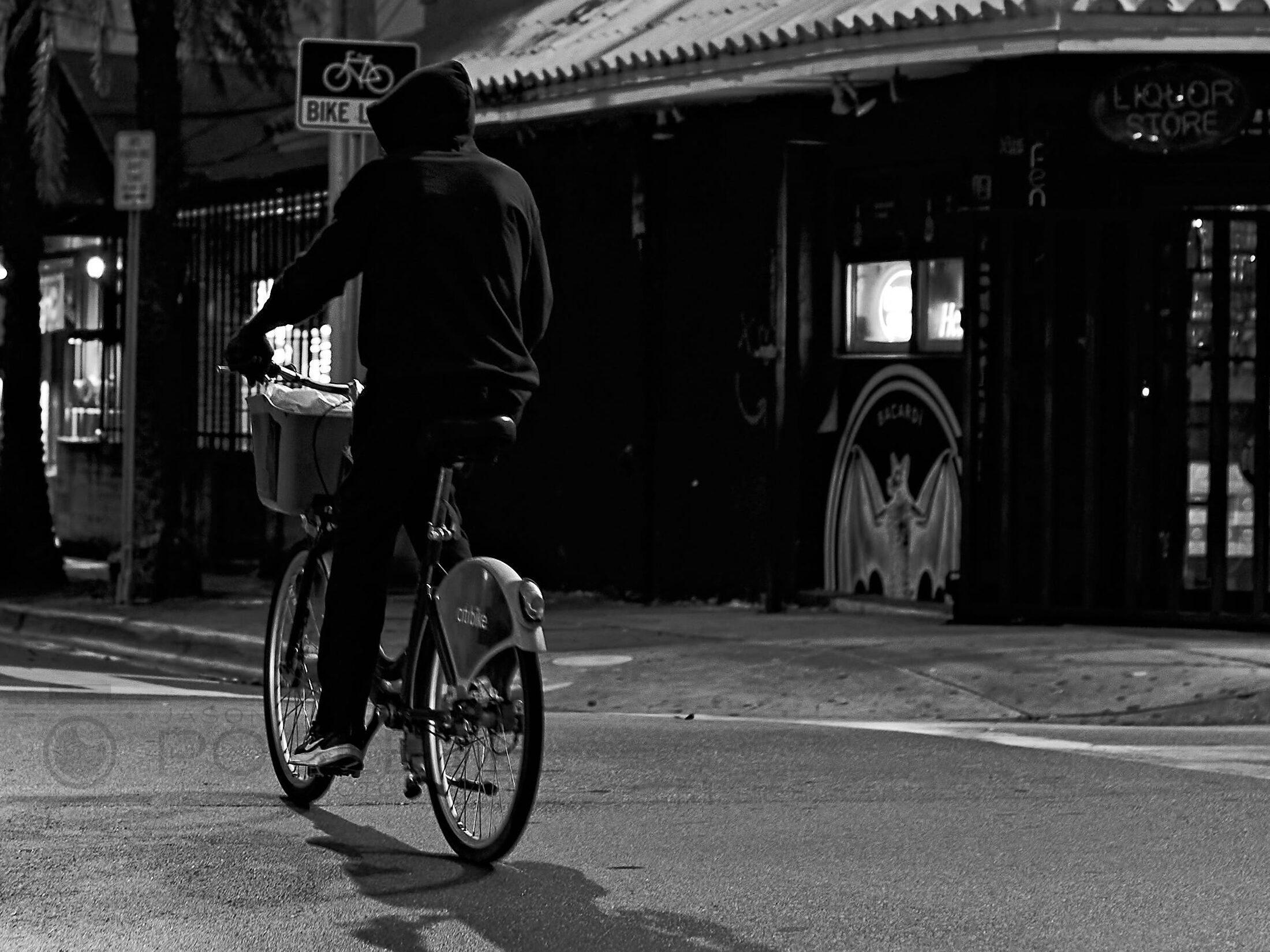 A cyclist crosses a sunlit street in front of dark storefronts.