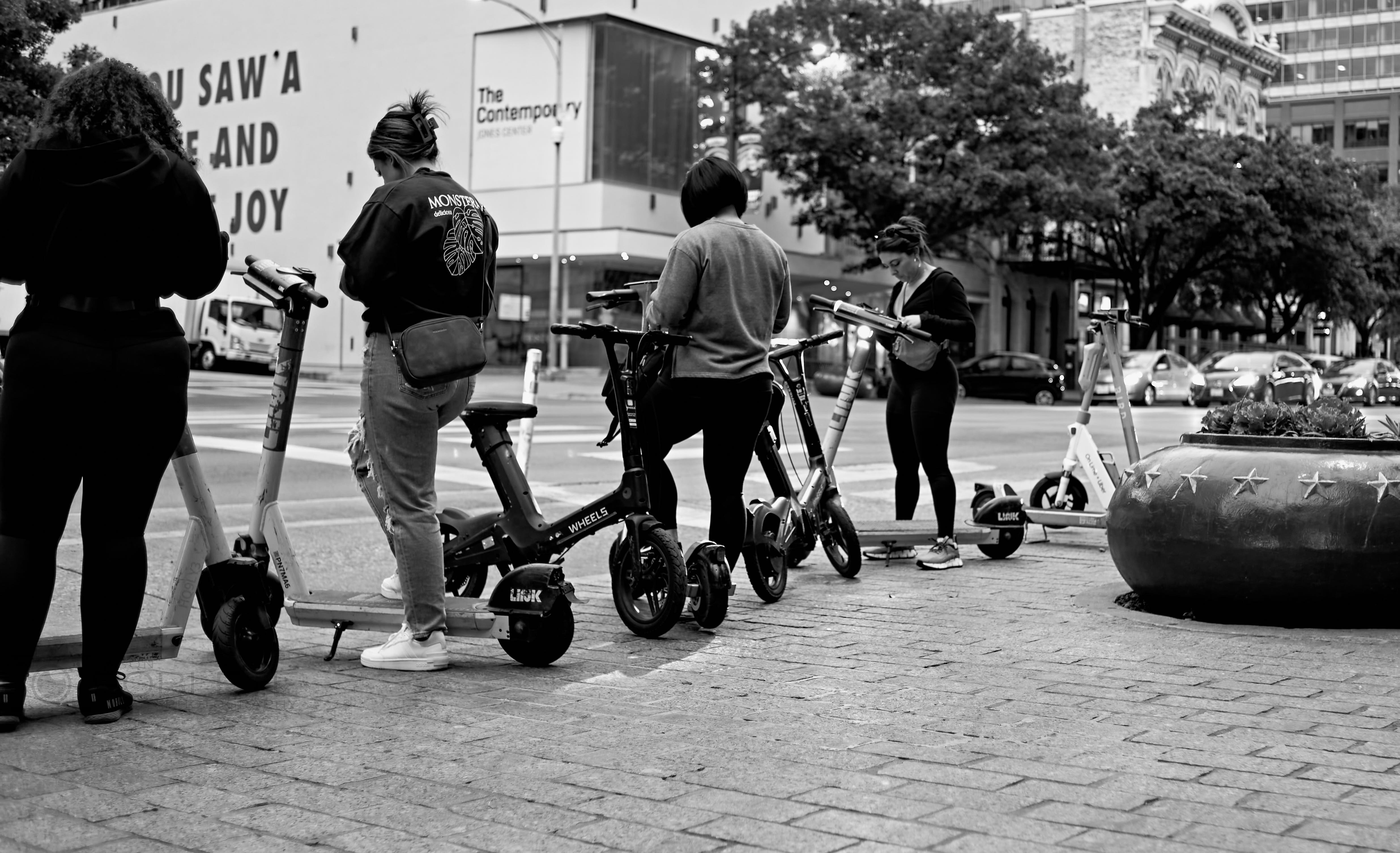 People gather with rental scooters on a downtown sidewalk.