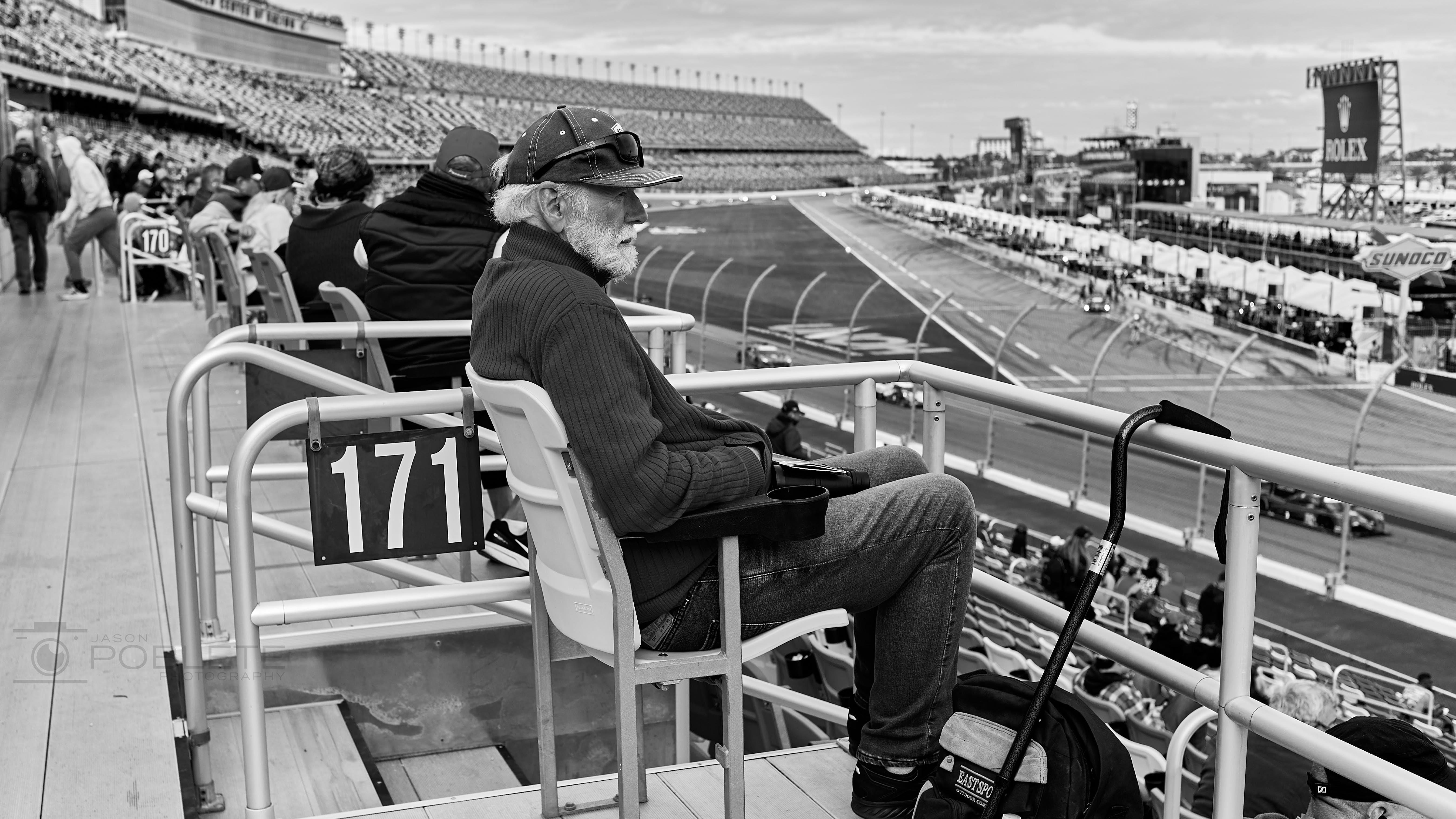 An older spectator sits above the track at a motor speedway.
