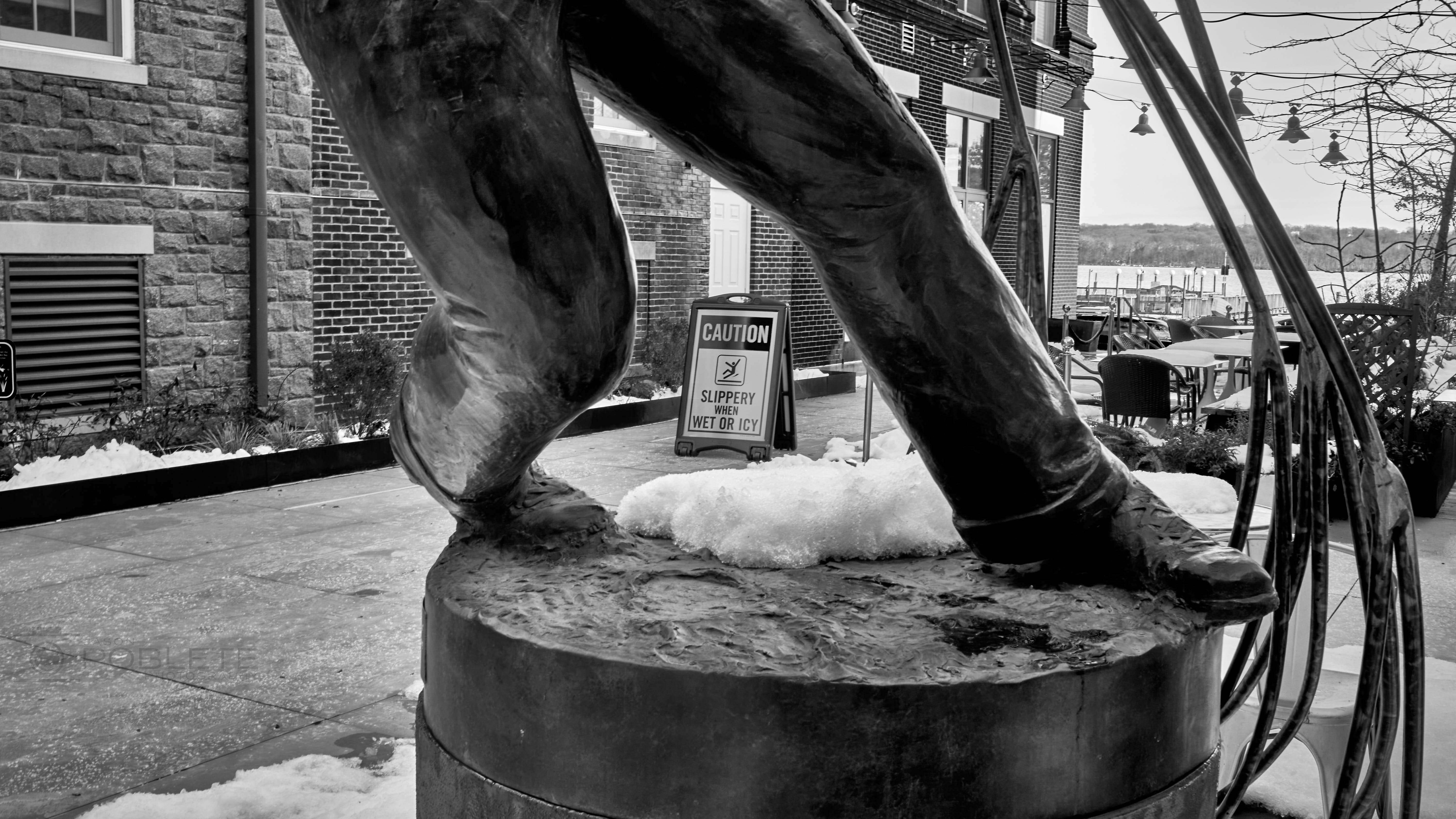A bronze statue frames a caution sign beside a snowy waterfront walkway.