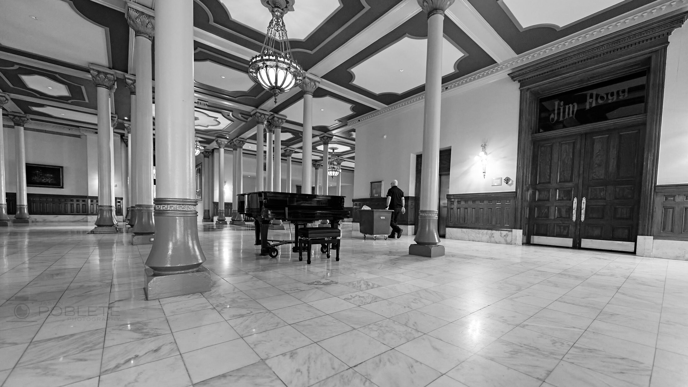 A worker pushes a cart past a grand piano in an ornate columned lobby.