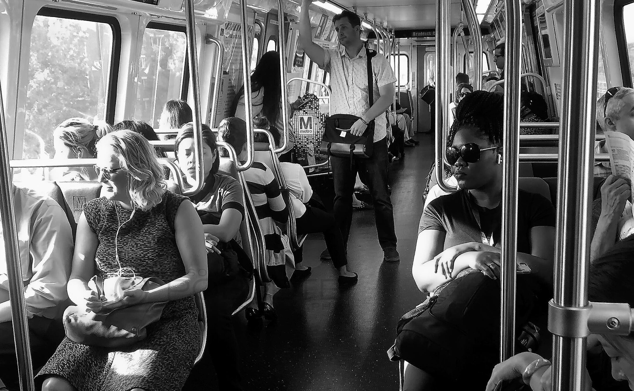Passengers sit and stand along the aisle of a sunlit city bus.