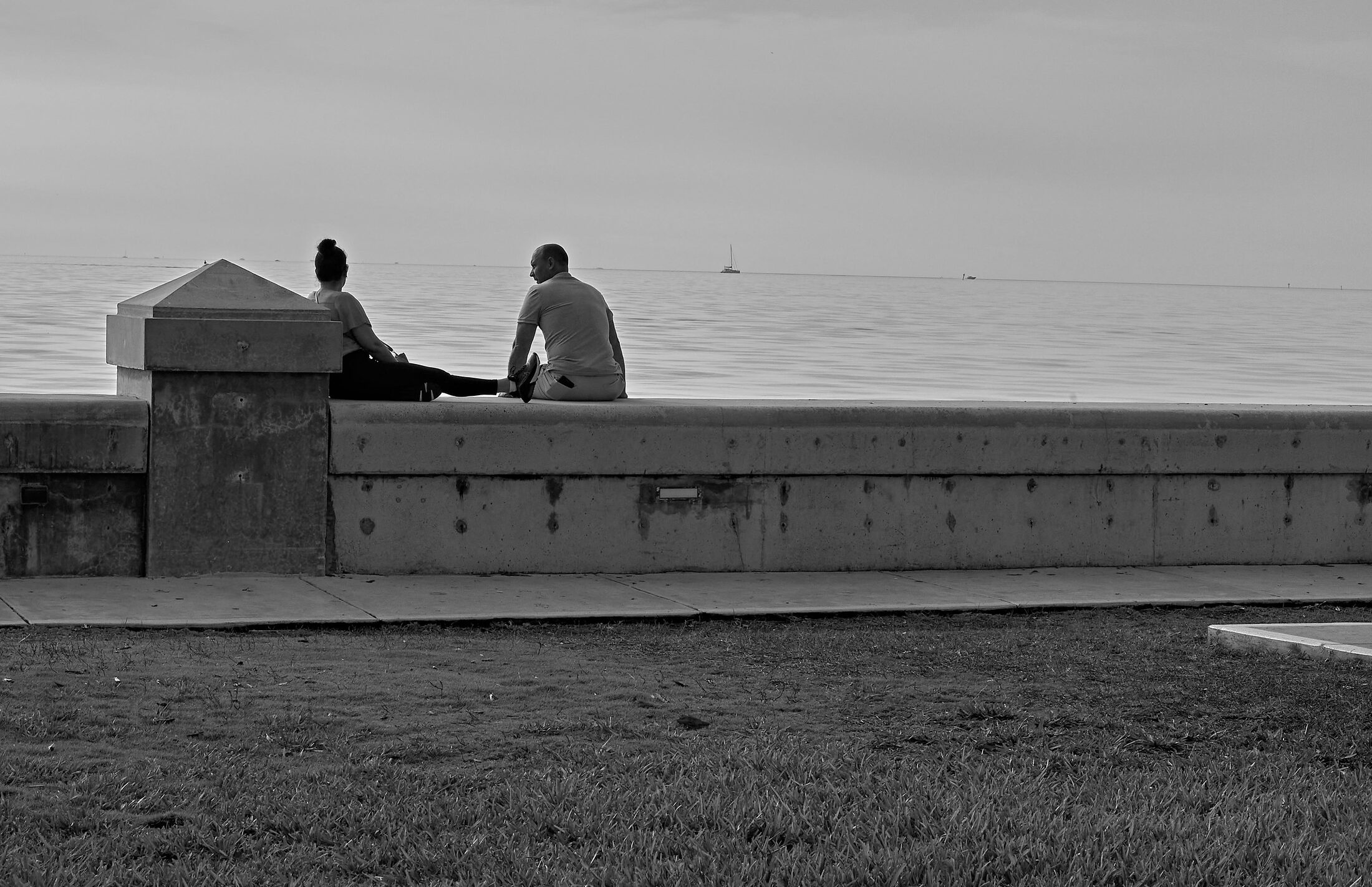 Two people sit on a seawall facing a calm horizon.