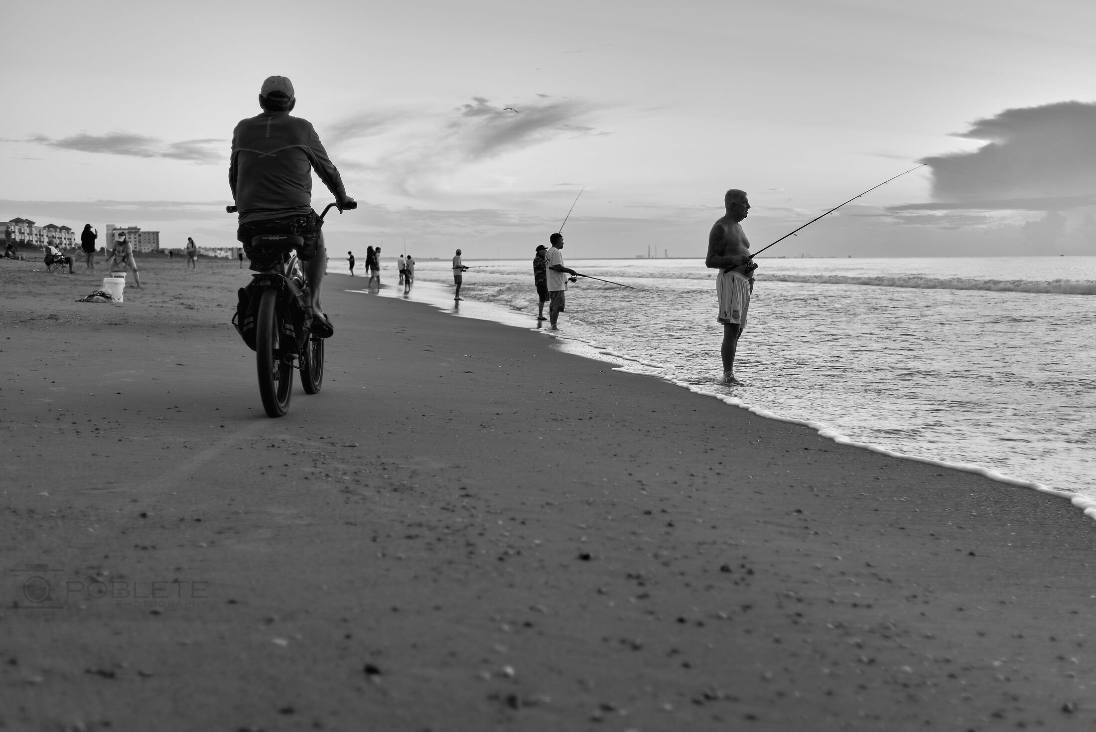 A cyclist rides along the shoreline as people fish from the beach.